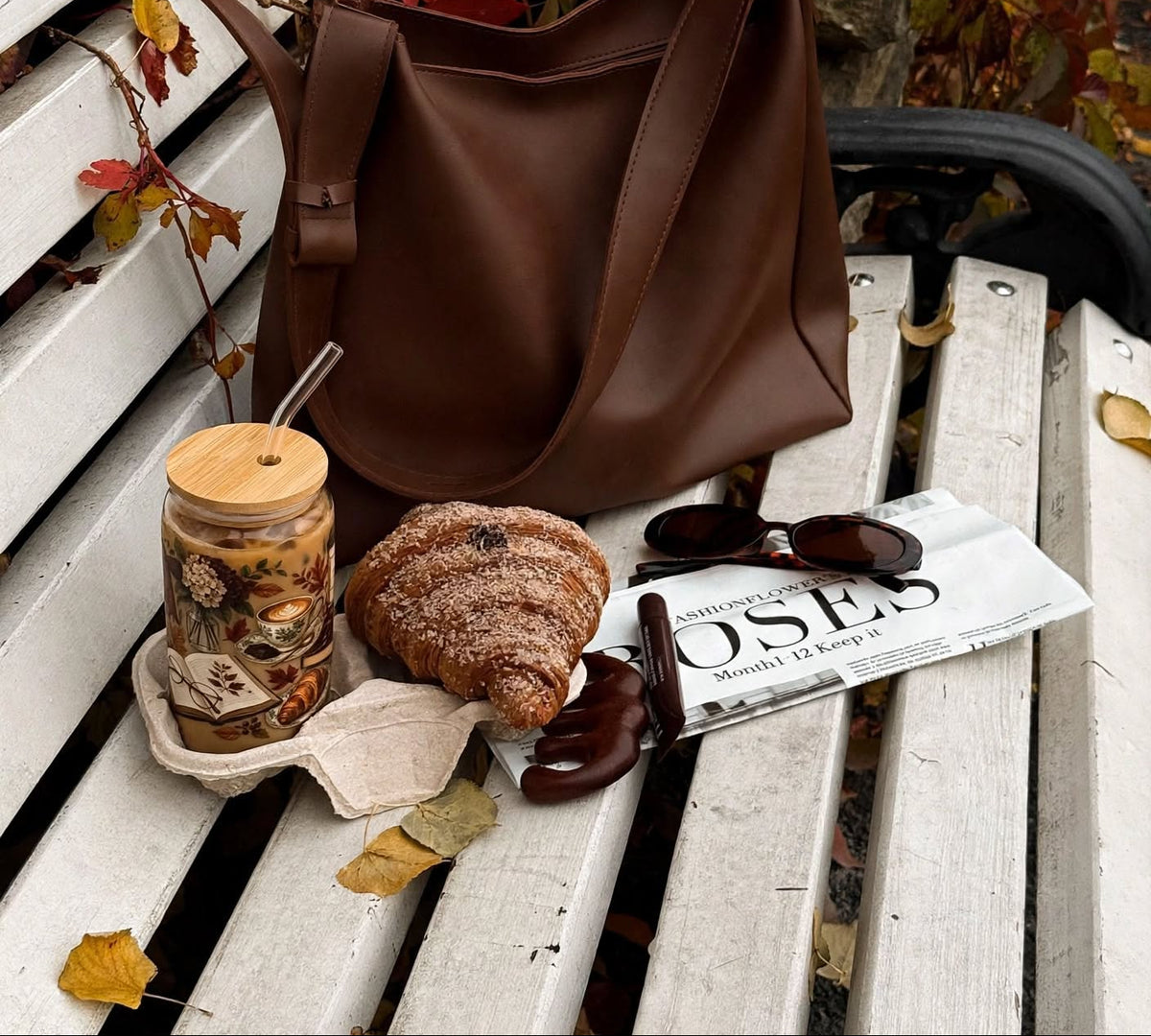 A brown leather shoulder bag sits on a white wooden bench outdoors, styled with a jar of coffee, a croissant, sunglasses, and a magazine in an autumn setting with red leaves.