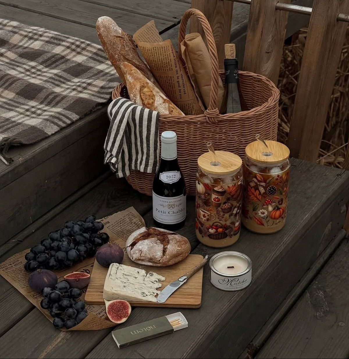 A styled picnic setup on a wooden deck featuring a wicker basket with bread and a bottle, two patterned glass jars with bamboo lids, a bottle of wine, cheese, grapes, figs, and a candle arranged on a table runner.